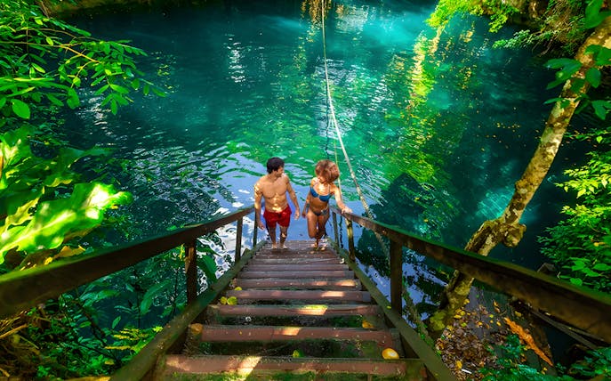 Couple ascending wooden stairs from a cenote surrounded by lush greenery.