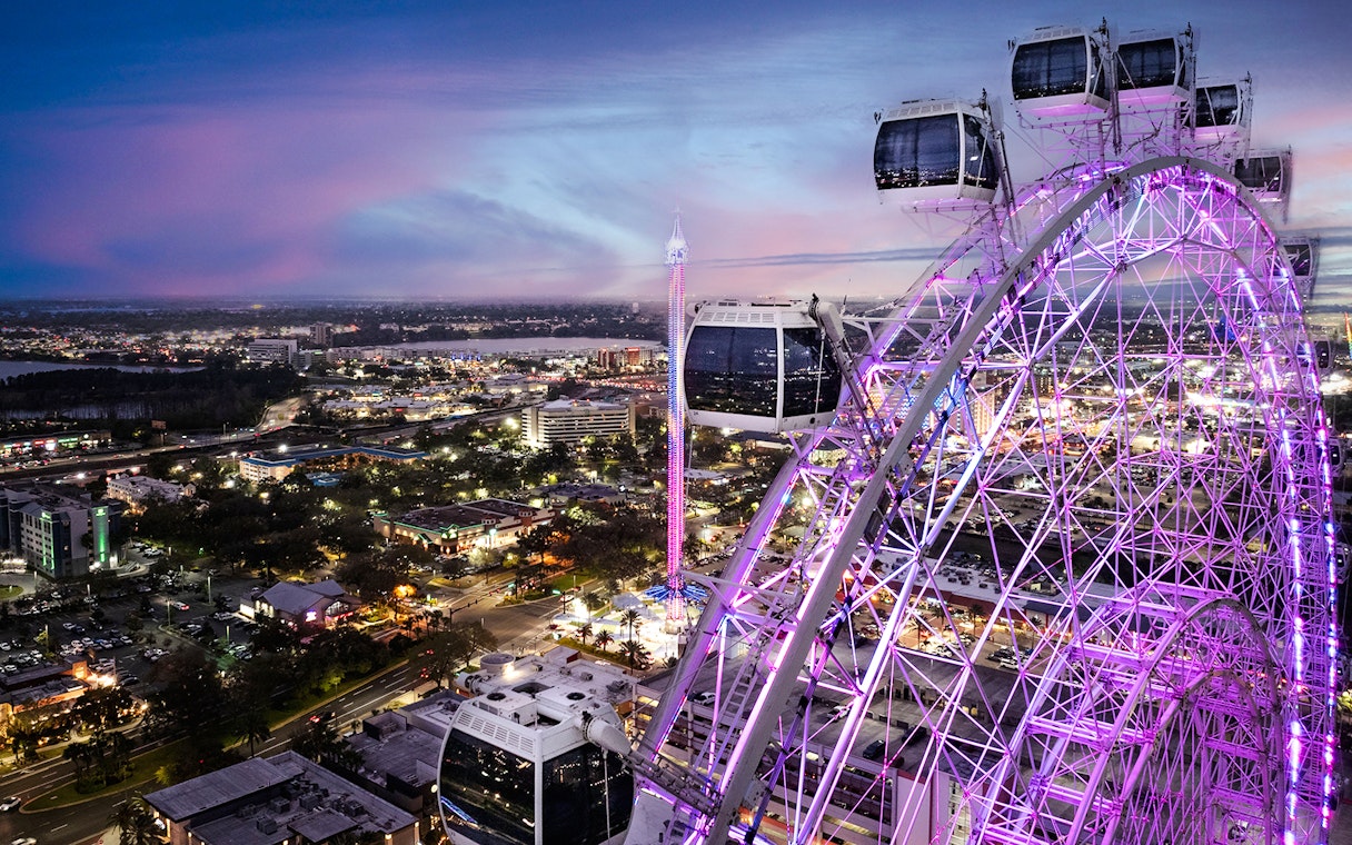 Ferris wheel at ICON Park, Orlando, illuminated at dusk with cityscape in the background.