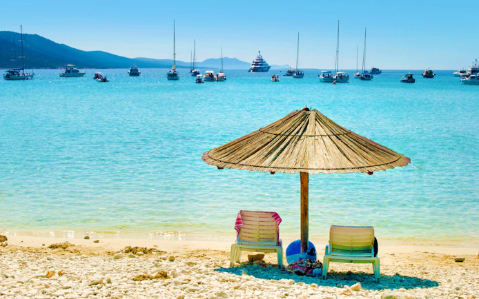 Sun loungers under a straw umbrella on Sakarun Beach, Croatia, with boats in the distance.