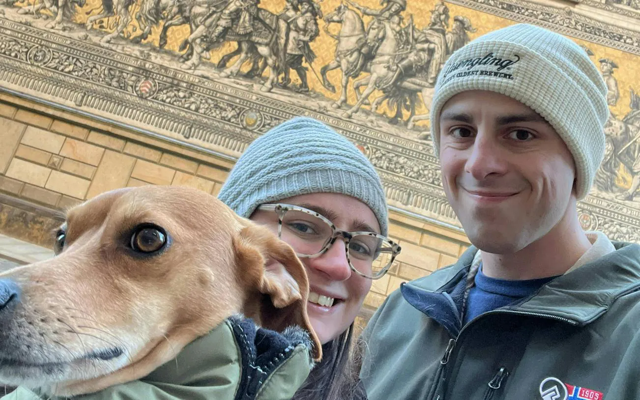 Couple with dog in front of Dresden's Fürstenzug mural during city exploration game.