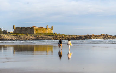 Two people walking on Matosinhos beach with a historic fort in the background, Porto.