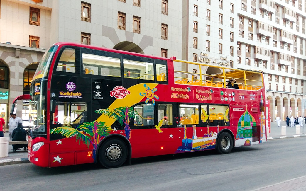 Red double-decker bus for Al Madinah Hop-On Hop-Off Tour parked near city buildings.