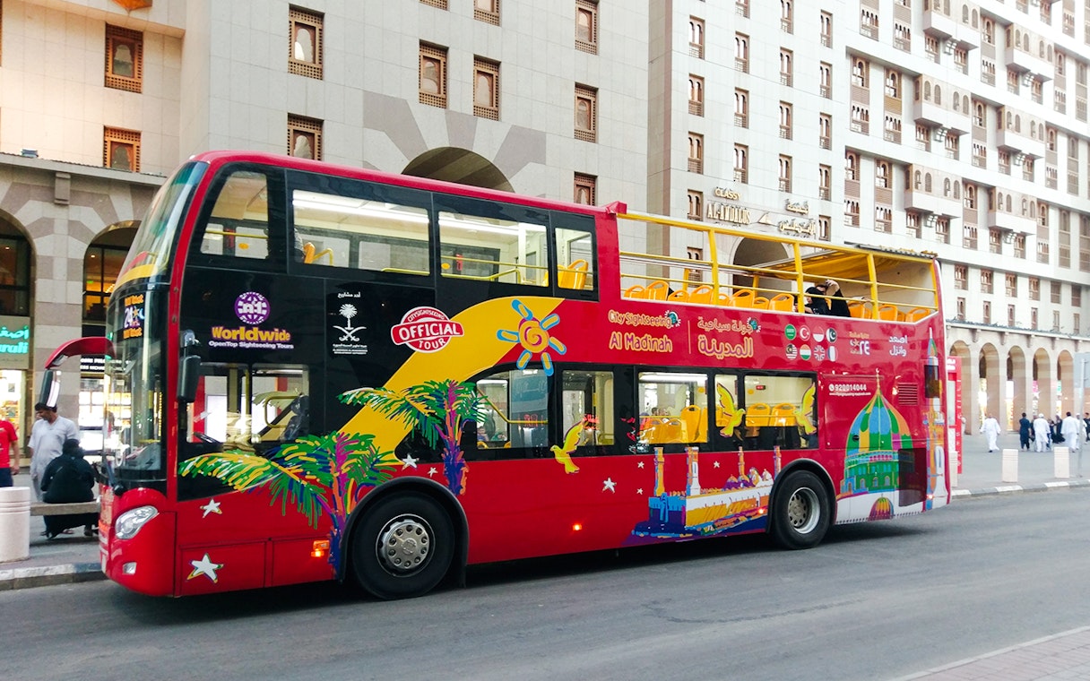 Red double-decker bus for Al Madinah Hop-On Hop-Off Tour parked near city buildings.