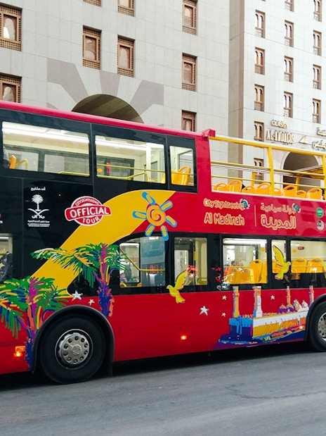 Red double-decker bus for Al Madinah Hop-On Hop-Off Tour parked near city buildings.