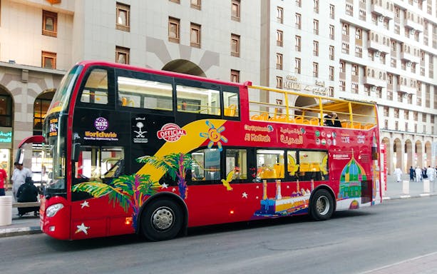 Red double-decker bus for Al Madinah Hop-On Hop-Off Tour parked near city buildings.