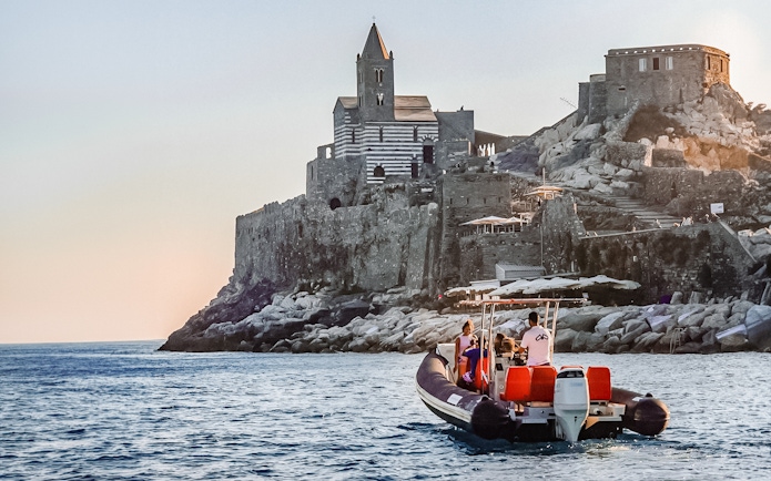 Boat tour approaching historic church on rocky coast, Cinque Terre, Italy.