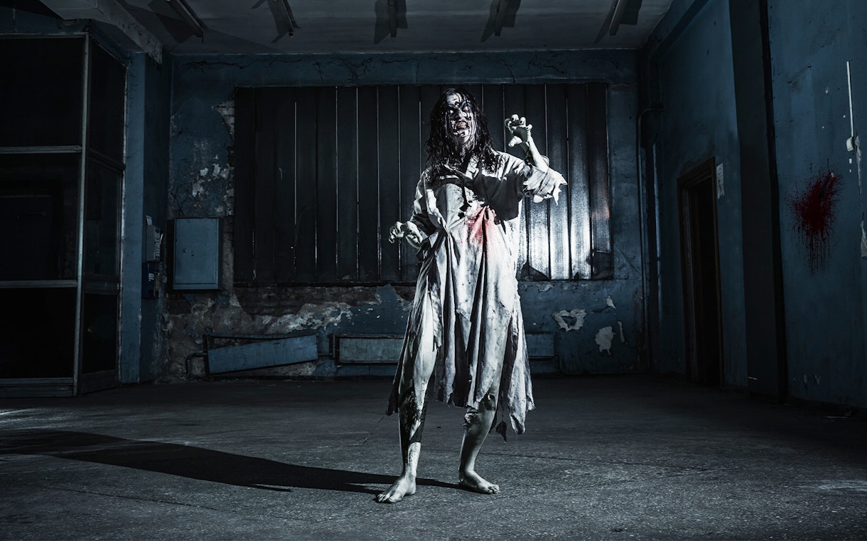 Zombie woman in tattered dress inside a dimly lit, abandoned haunted house.
