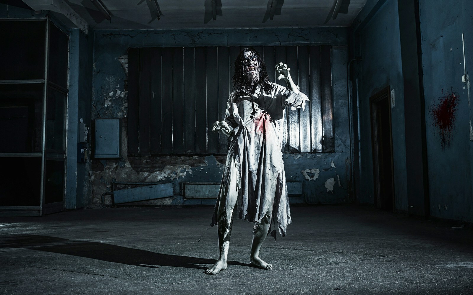 Zombie woman in tattered dress inside a dimly lit, abandoned haunted house.