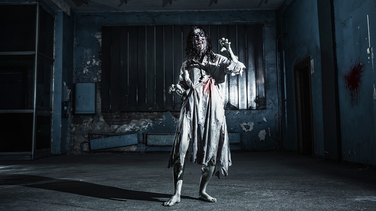 Zombie woman in tattered dress inside a dimly lit, abandoned haunted house.