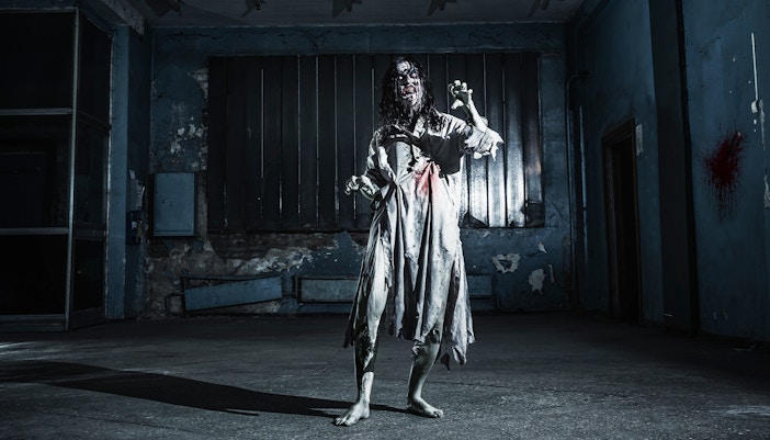 Zombie woman in tattered dress inside a dimly lit, abandoned haunted house.