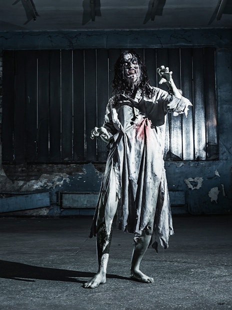 Zombie woman in tattered dress inside a dimly lit, abandoned haunted house.