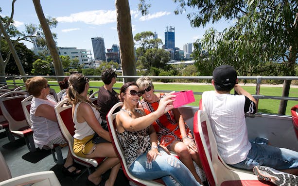 Tourists on a hop-on-hop-off bus in Kings Park, Perth, with city skyline in the background.
