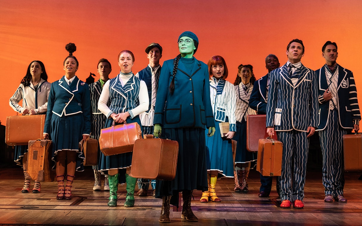 Cast of "Wicked" in school uniforms holding suitcases on stage.