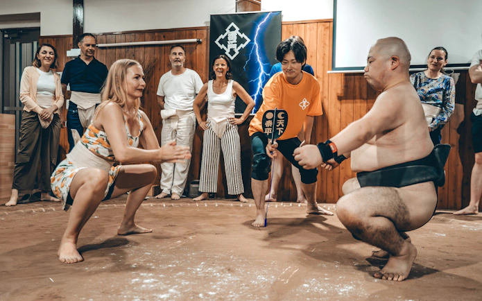Woman in athletic wear and sumo wrestler squatting in a ring, observed by a referee and spectators.