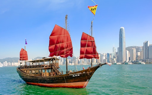 AquaLuna junk boat with red sails cruising Victoria Harbour, Hong Kong skyline in background.