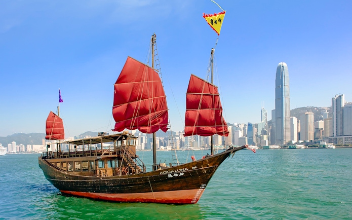 AquaLuna junk boat with red sails cruising Victoria Harbour, Hong Kong skyline in background.