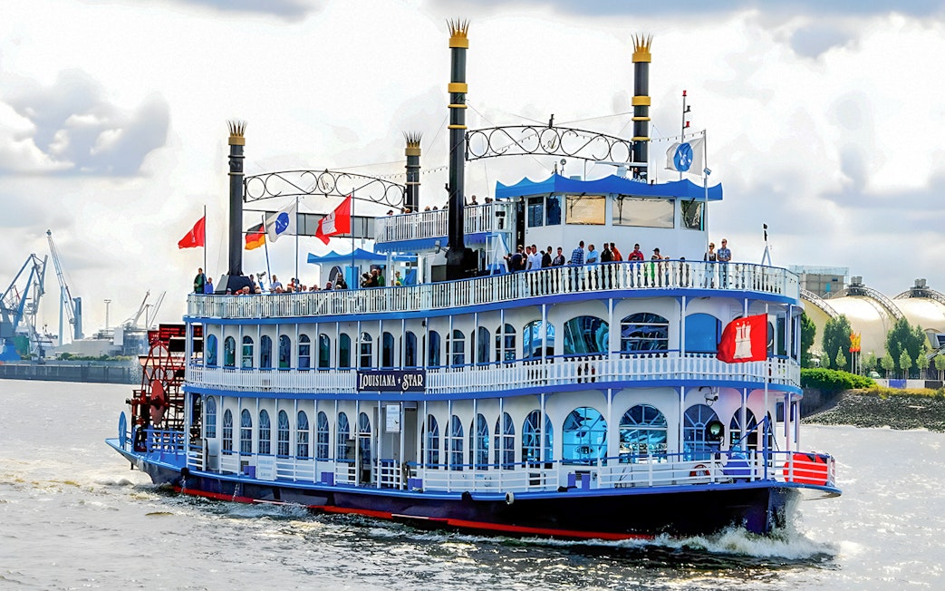 Paddle steamer on Hamburg Harbor cruise with passengers on deck, cityscape in background.