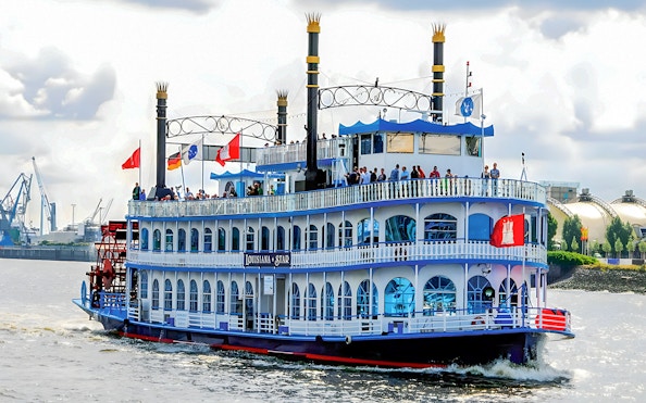 Paddle steamer on Hamburg Harbor cruise with passengers on deck, cityscape in background.