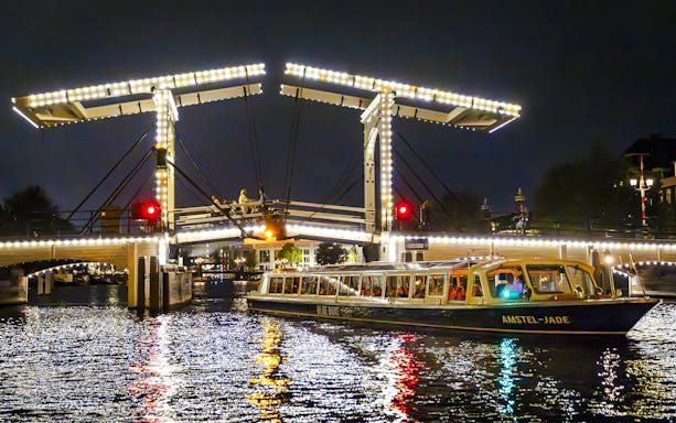 Evening cruise boat passing under illuminated bridge in Amsterdam.