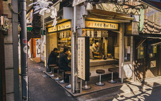 Tokyo street ramen shop with customers seated at a counter, chef preparing dishes.