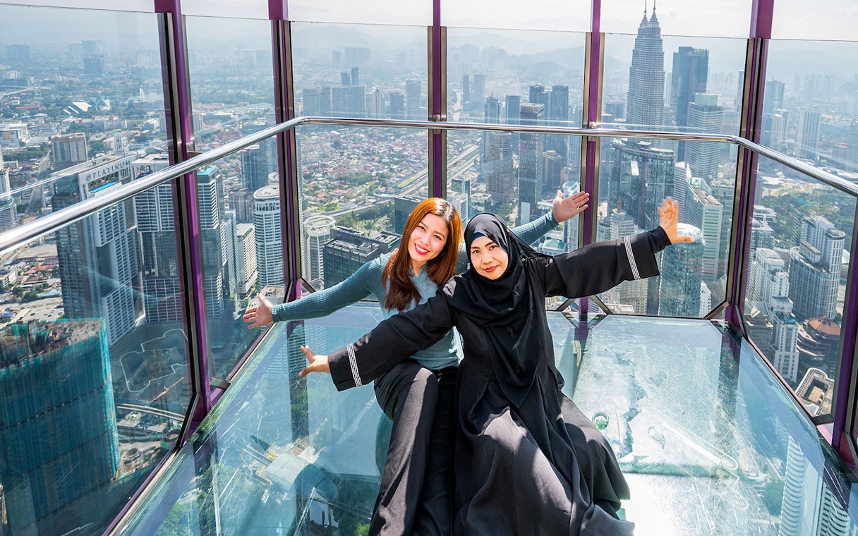 Tourists posing in the Kuala Lumpur Tower Glass Box with city skyline view.