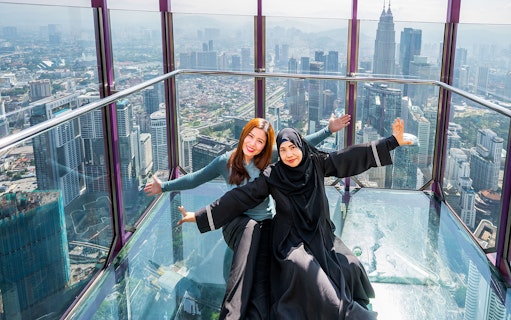 Tourists posing in the Kuala Lumpur Tower Glass Box with city skyline view.