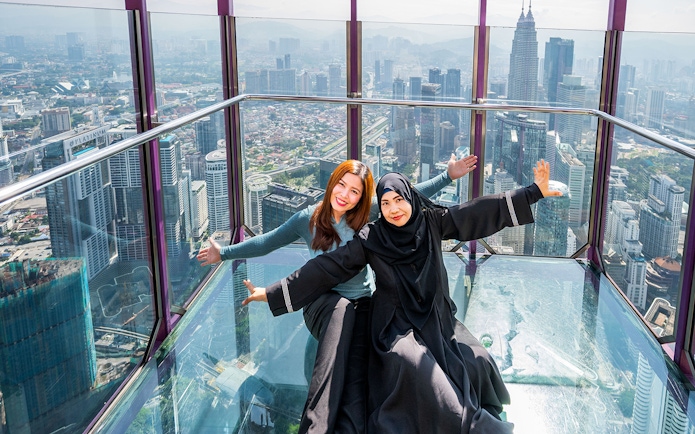Tourists posing in the Kuala Lumpur Tower Glass Box with city skyline view.
