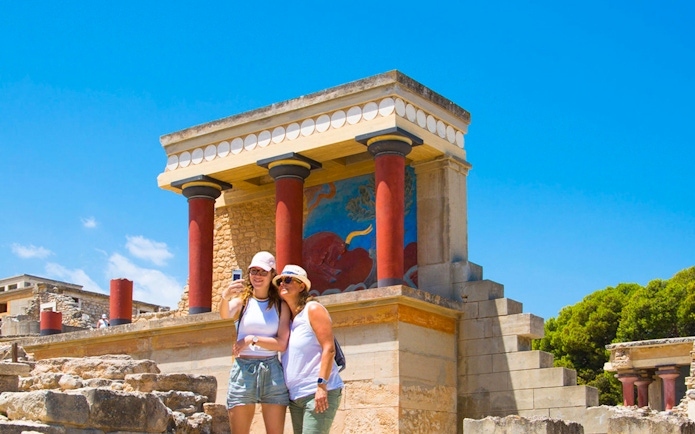 Visitors taking a selfie at Knossos Palace, Crete, with ancient fresco and columns in view.