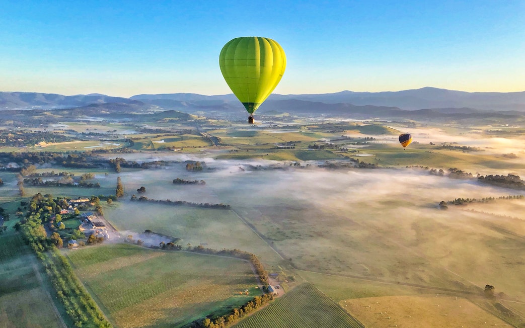 Hot air balloons floating over Yarra Valley at sunrise, with misty fields and distant hills.