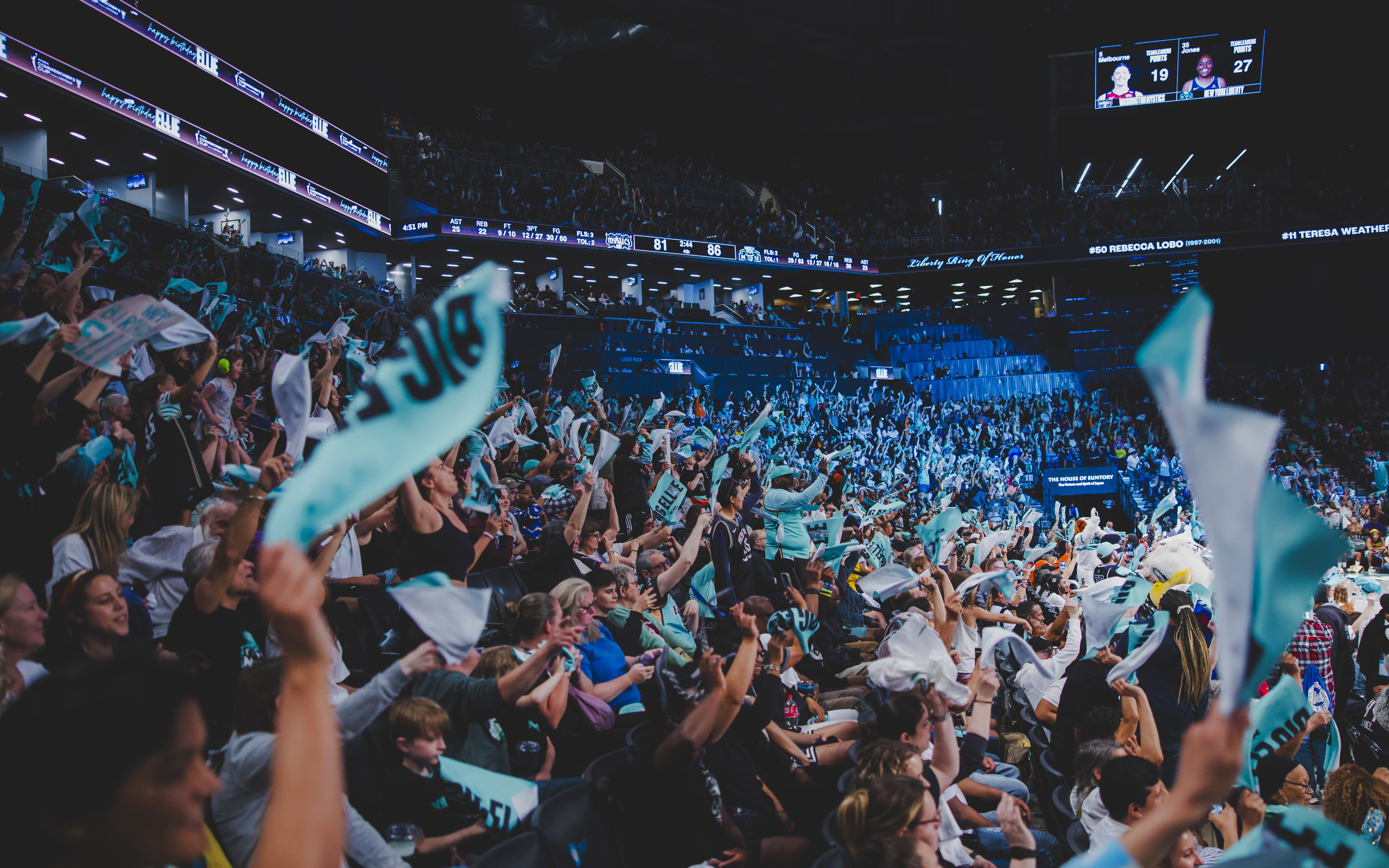 Fans cheering at New York Liberty basketball game in a packed arena.