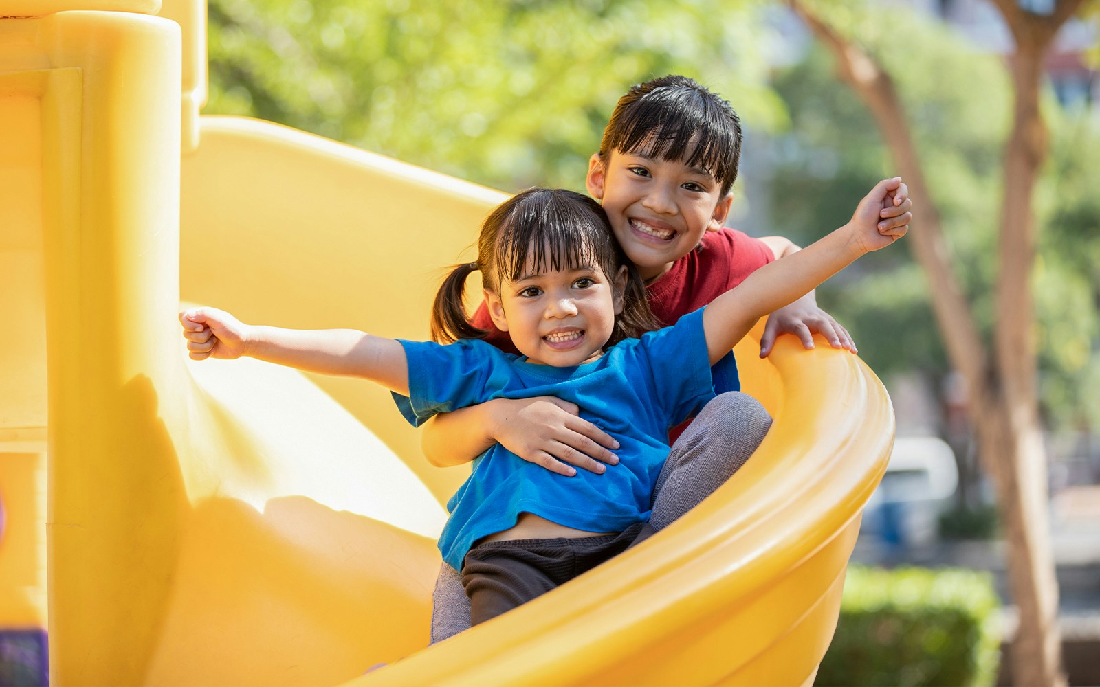 Children sliding down a yellow slide in a garden play area.