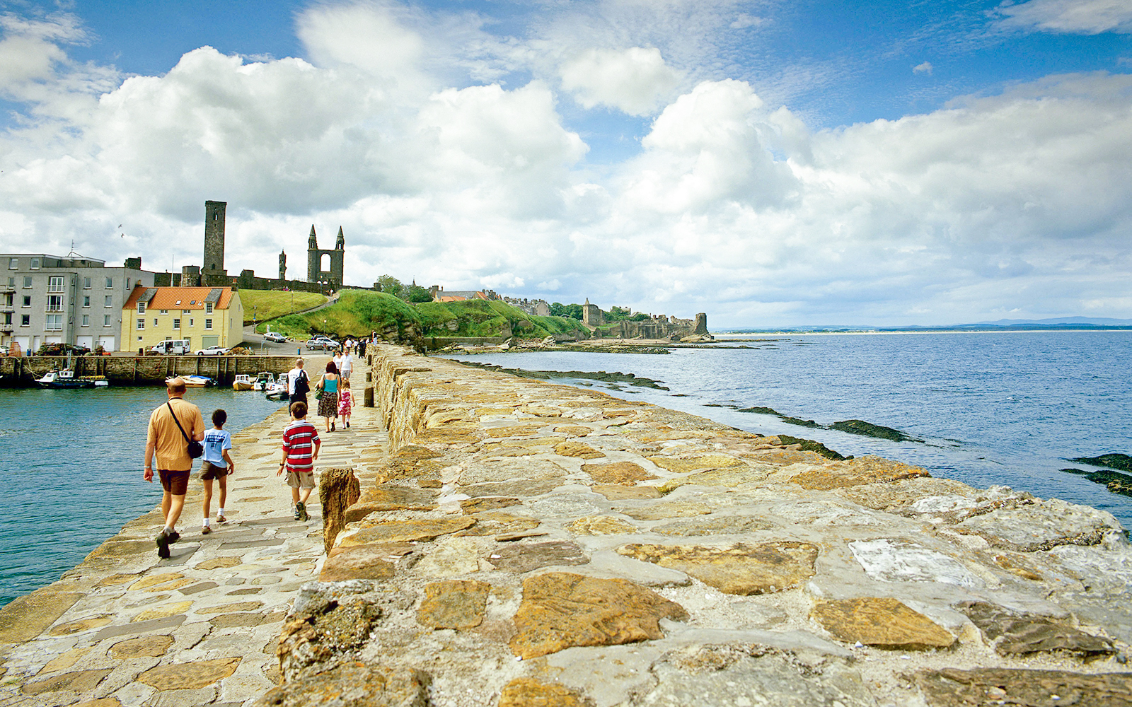 Tourists walking on coastal path with St. Andrews Cathedral ruins in background.