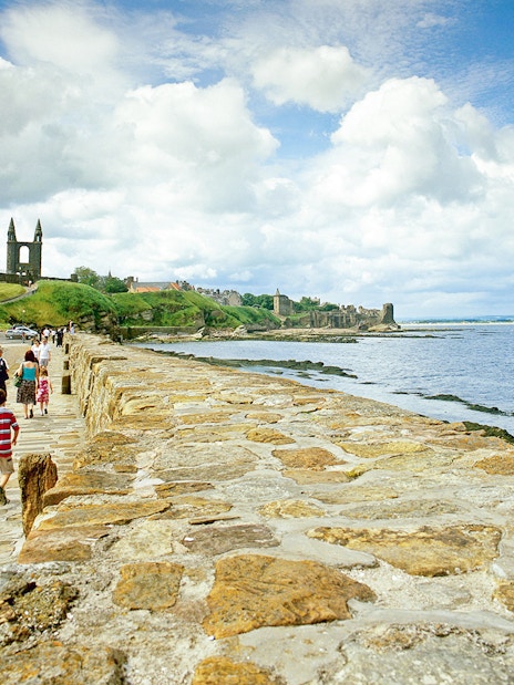 Tourists walking on coastal path with St. Andrews Cathedral ruins in background.