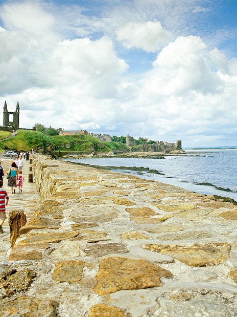 Tourists walking on coastal path with St. Andrews Cathedral ruins in background.