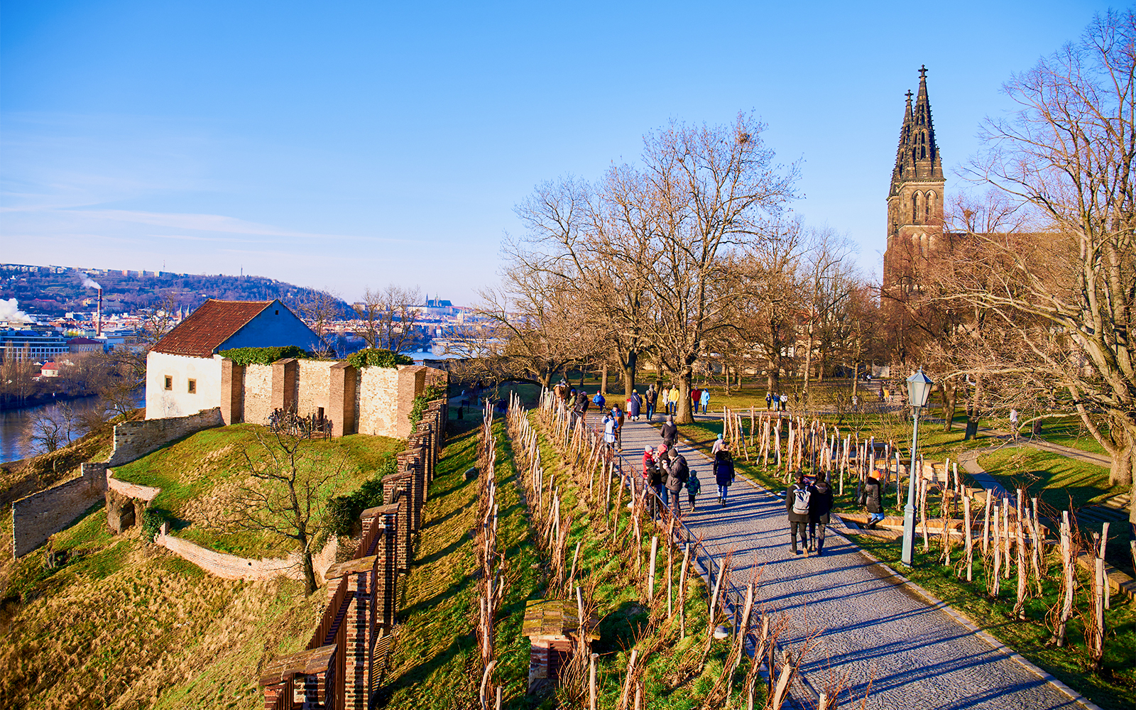 Vyšehrad park path with visitors, historic church tower, and scenic city view in Prague.