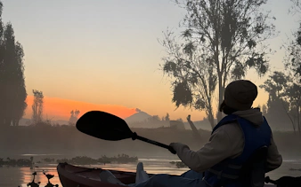 Kayaking at sunrise in Xochimilco canals with silhouetted trees and mist.