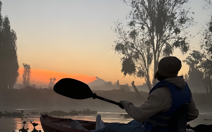 Kayaking at sunrise in Xochimilco canals with silhouetted trees and mist.