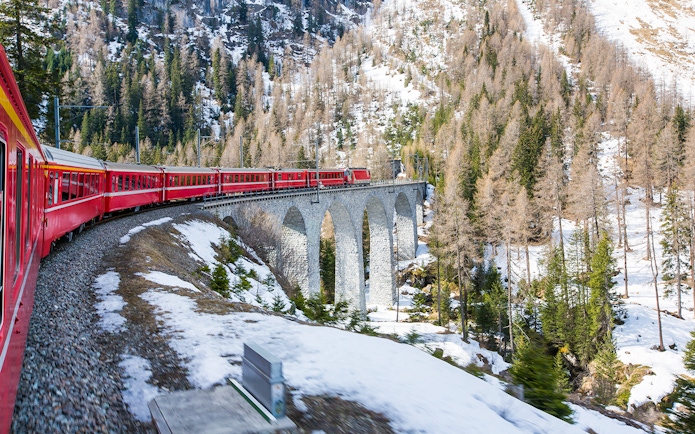 Bernina train crossing a stone viaduct in a snowy alpine landscape.