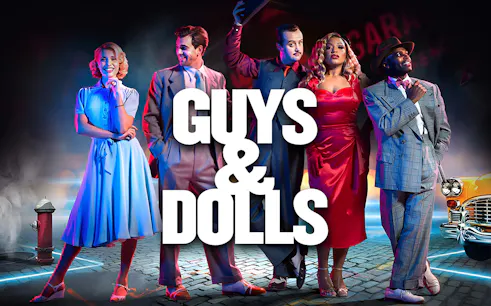 Cast of Guys & Dolls in vintage attire on a city street with neon lights.