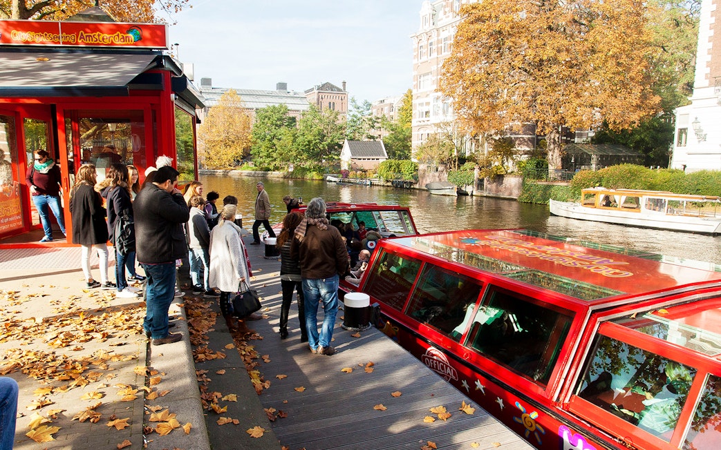 Tourists boarding a red canal boat for a City Sightseeing hop-on hop-off tour in Amsterdam.