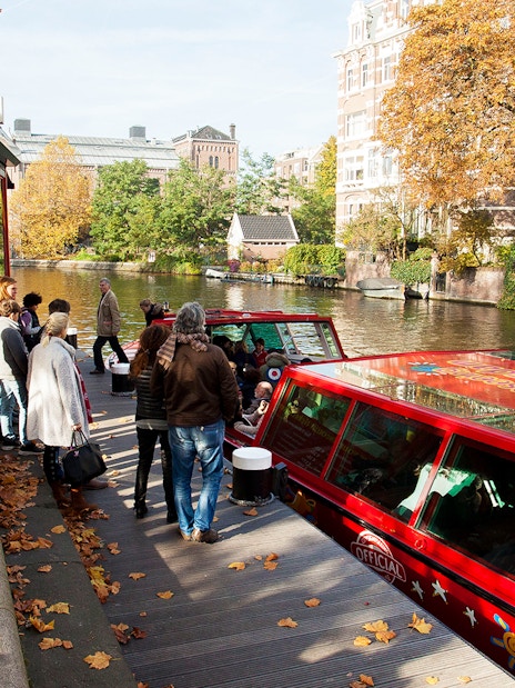 Tourists boarding a red canal boat for a City Sightseeing hop-on hop-off tour in Amsterdam.