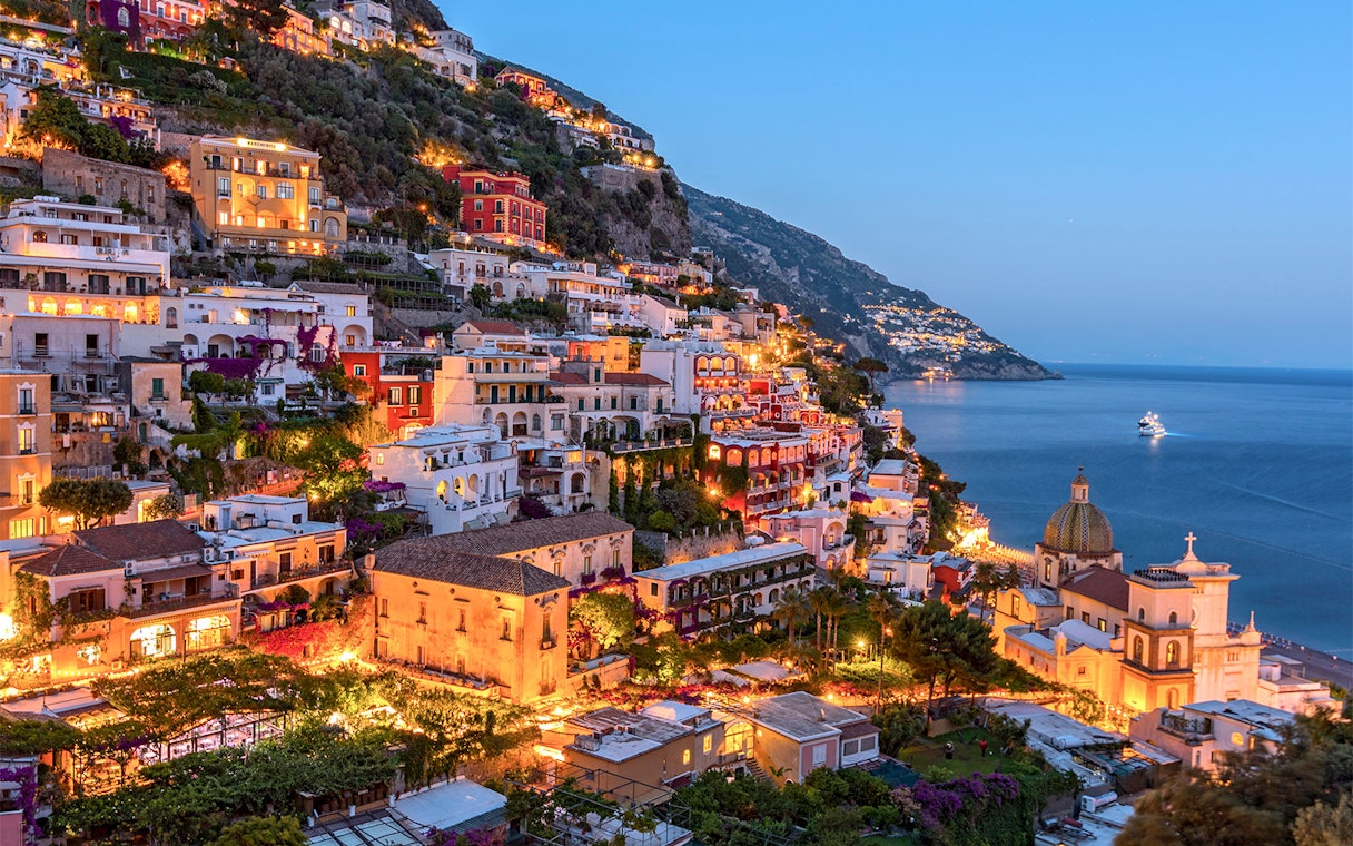 Amalfi Coast hillside view with illuminated buildings and sea, part of Naples day tour.