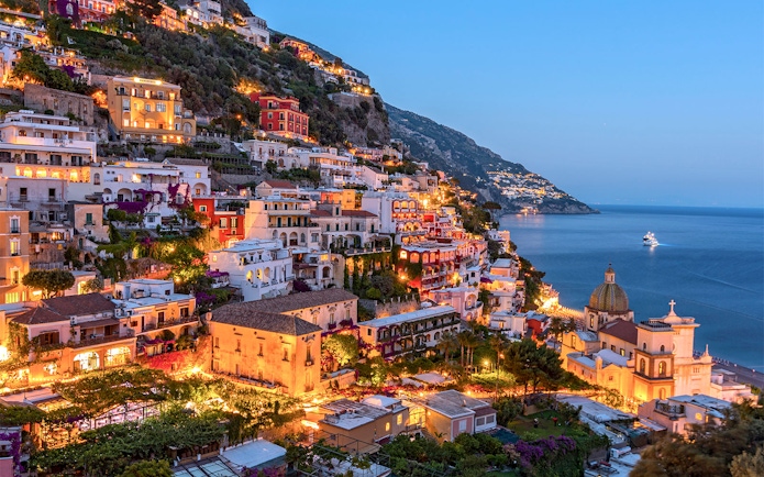 Amalfi Coast hillside view with illuminated buildings and sea, part of Naples day tour.