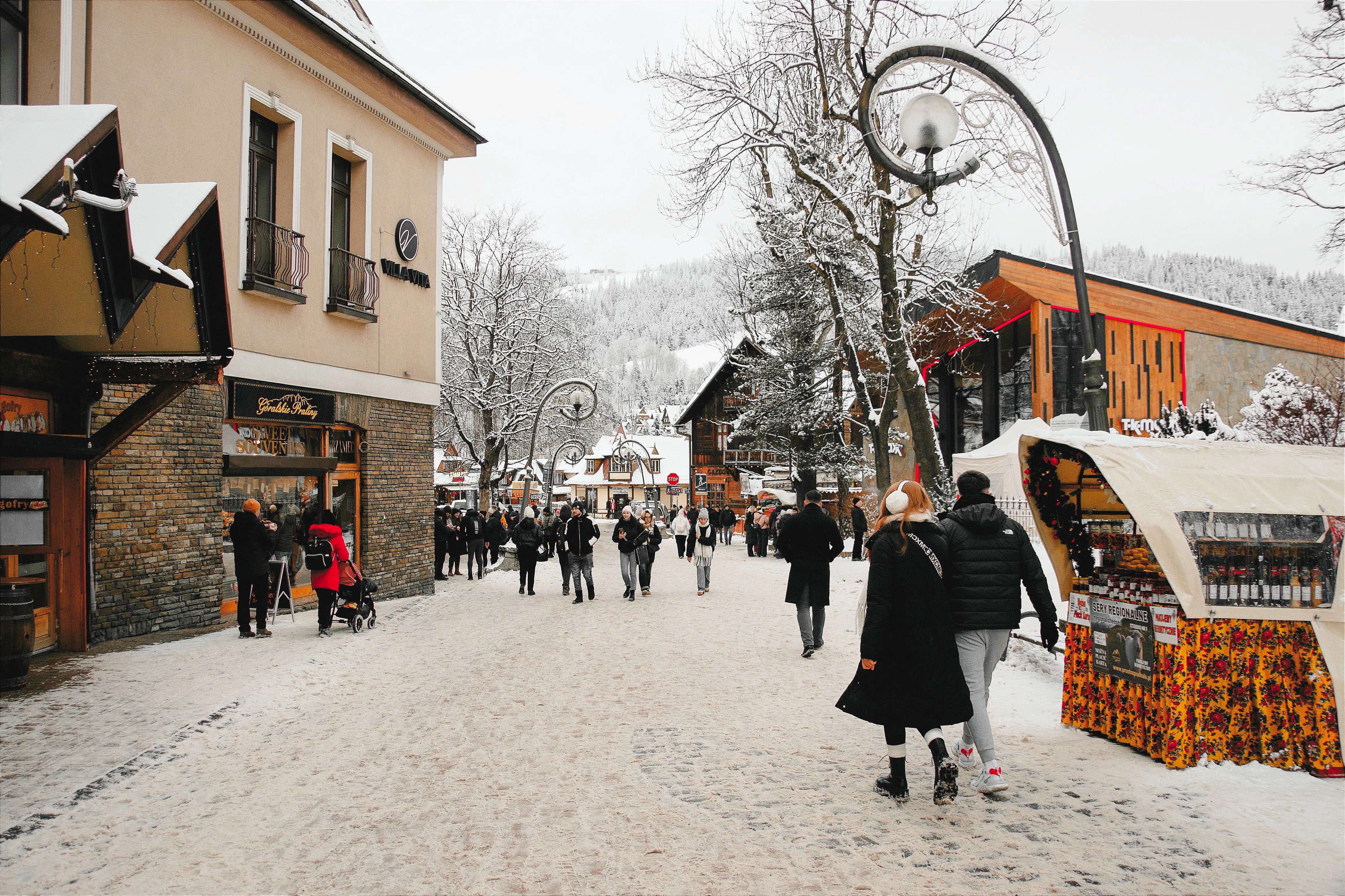 Pedestrians walking on a snowy street in Zakopane, Poland, with shops and snow-covered trees.