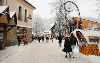 Pedestrians walking on a snowy street in Zakopane, Poland, with shops and snow-covered trees.