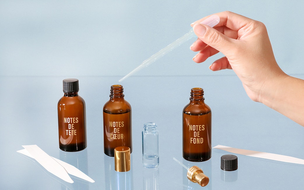 Hand using dropper with perfume bottles labeled "Notes de Tête," "Notes de Cœur," "Notes de Fond" in Grasse workshop.