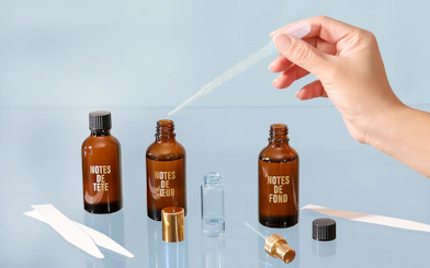 Hand using dropper with perfume bottles labeled "Notes de Tête," "Notes de Cœur," "Notes de Fond" in Grasse workshop.