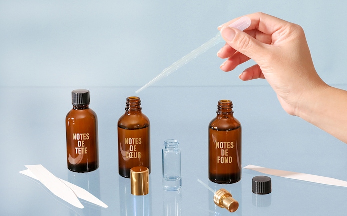 Hand using dropper with perfume bottles labeled "Notes de Tête," "Notes de Cœur," "Notes de Fond" in Grasse workshop.