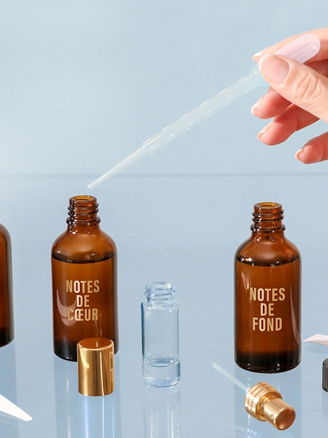 Hand using dropper with perfume bottles labeled "Notes de Tête," "Notes de Cœur," "Notes de Fond" in Grasse workshop.