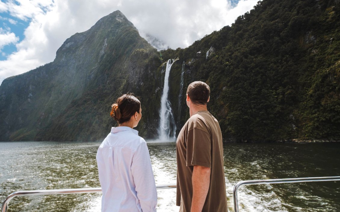 Couple on Milford Sound cruise viewing waterfall and lush mountains.